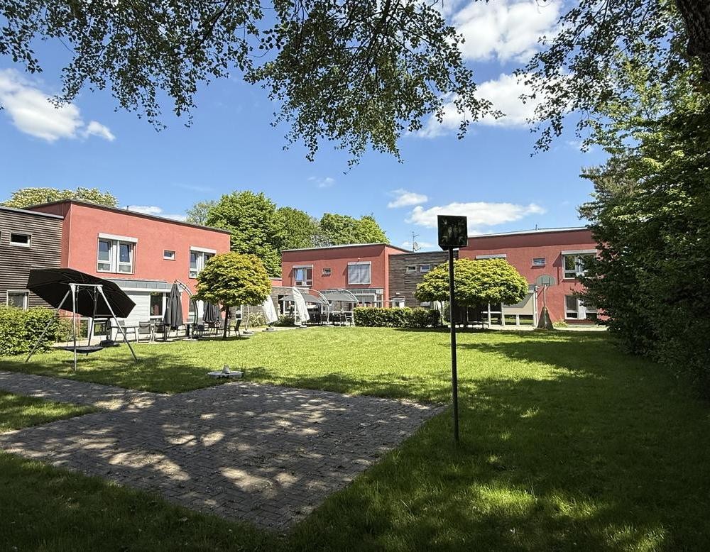 Gartenansicht eines Hauses mit roter und brauner Fassade vielen Fenstern. Auf einer grünen Wiese steht ein Strandkorb mit gelb-graue gestreiftem Dach