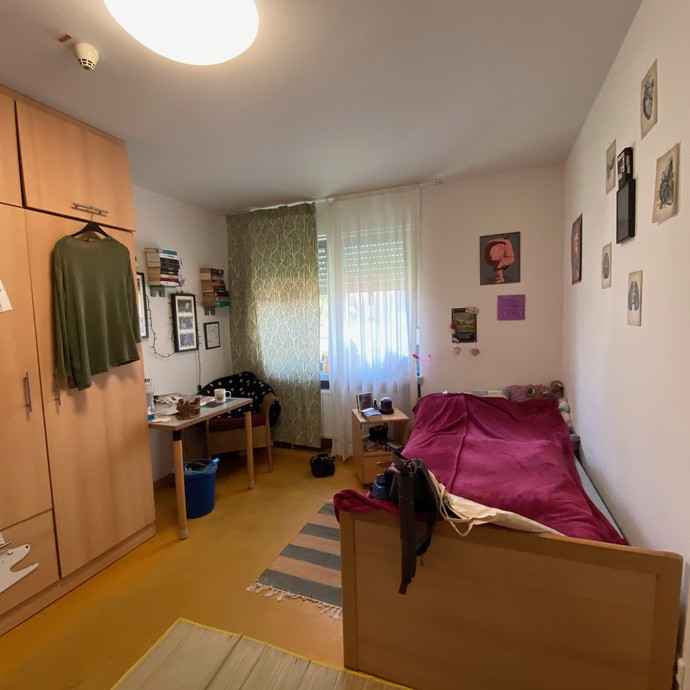 Ein Zimmer, rechts ein Bett mit roter Decke bezogen, links vor einem Fenster ein kleiner Schreibtisch (öffnet vergrößerte Bildansicht)
