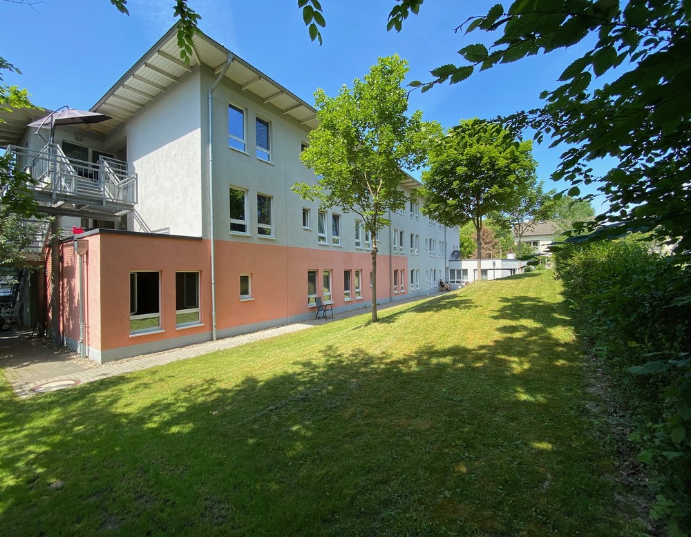 EinHaus mit grau-rosafarbiger Fassade. Im Vordergrund schattenspendende Bäume bei sonnigem Wetter