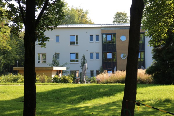 Zwischen zwei Baumstämmen erkenn man ein dreistöckiges Gebäude mit weißer u d Gelder Fassade. Davor ein gepflegter Garten.