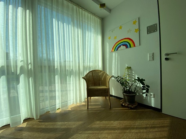 Ein Korbstuhl steht vor einem großflächigem Fenster mit einer luftigen Gardine davor