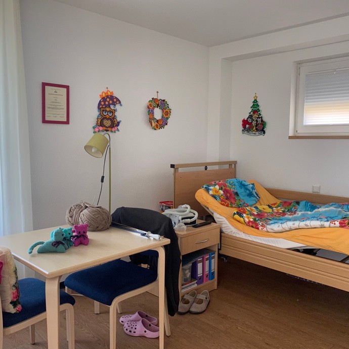 Blick in ein Zimmer mit Tisch im Vordergrund, dahinter ein mit bunter Bettwäsche bezogenes Bett (vergrößerte Bildansicht wird geöffnet)