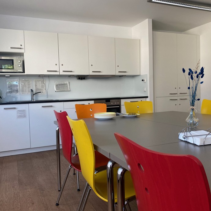 Bunte Stühle an einem Tisch mit weißer Tischplatte. Im Hinterrund einer moderne Küchenzeile (vergrößerte Bildansicht wird geöffnet)