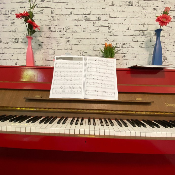 Ein rotes Klavier steht vor einer weißen Backsteinwand-Tapete,. Auf dem Klavier ein aufgeblättertes Notenheft (vergrößerte Bildansicht wird geöffnet)