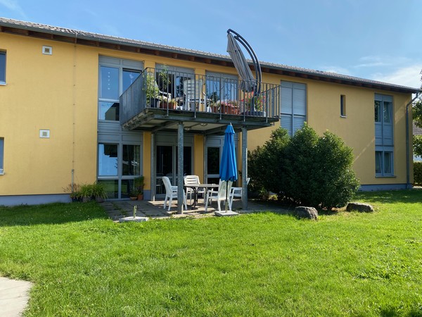 Ein Haus mit gelber Fassade und einem großzügigen Balkon. Darunter eine weiße Sitzgruppe und ein Sonnenschirm. Davor ein begrünter Garten