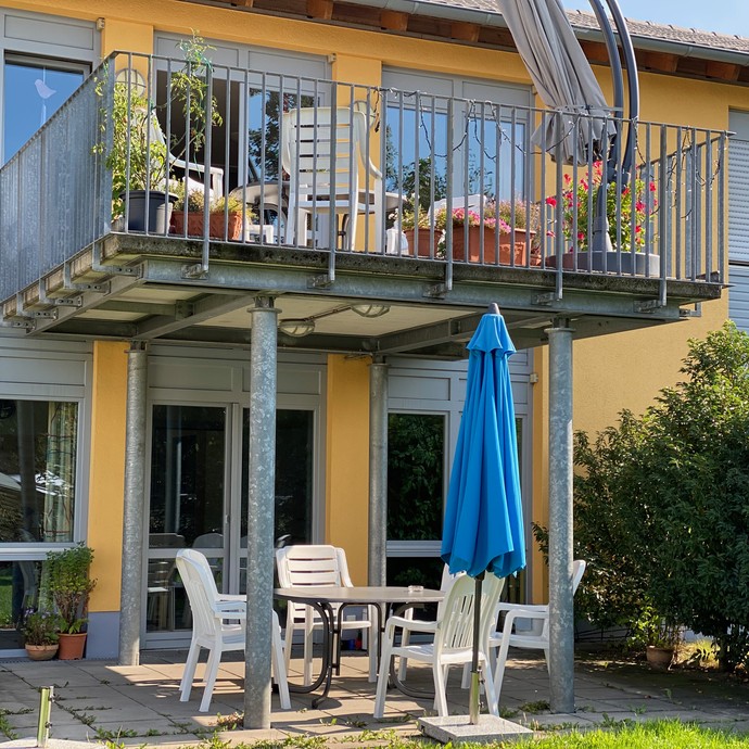 Ein Balkon mit Gartenmöbeln. Unter dem Balkon eine Sitzgruppe mit blauem Sonnenschirm (öffnet vergrößerte Bildansicht)