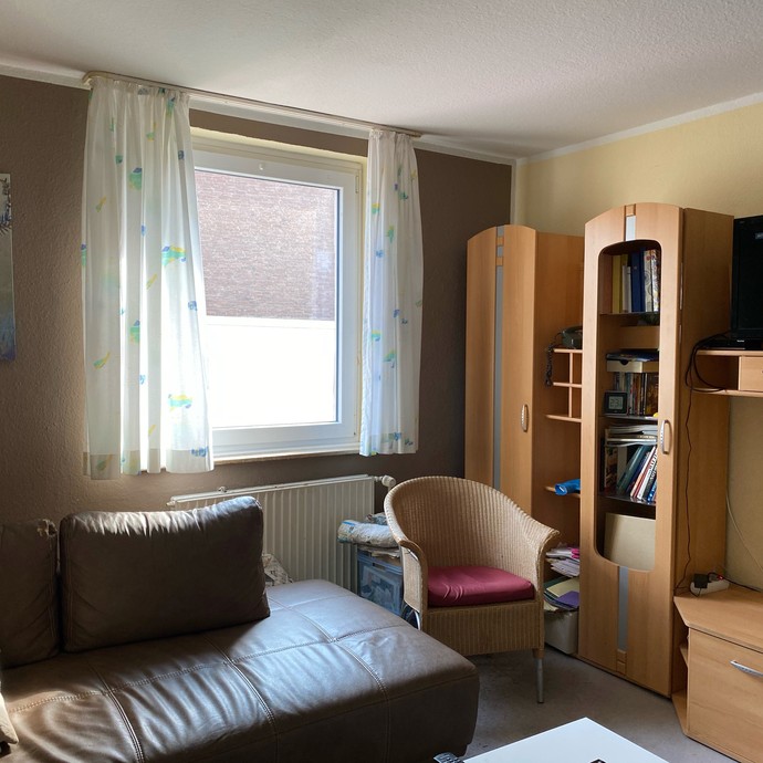 Blick in ein Zimmer, links steht eine graue Ledercouch, rechts steht eine Schrankwand mit hölzernem Furnier. Man blickt durch ein Fenster zum Garten (öffnet vergrößerte Bildansicht)