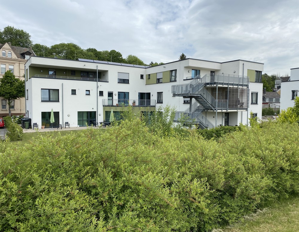 Hinter einer grünen Hecke steht ein modernes, weißes Gebäude mit Terrassen und eine Stahltreppe vor der Fassade