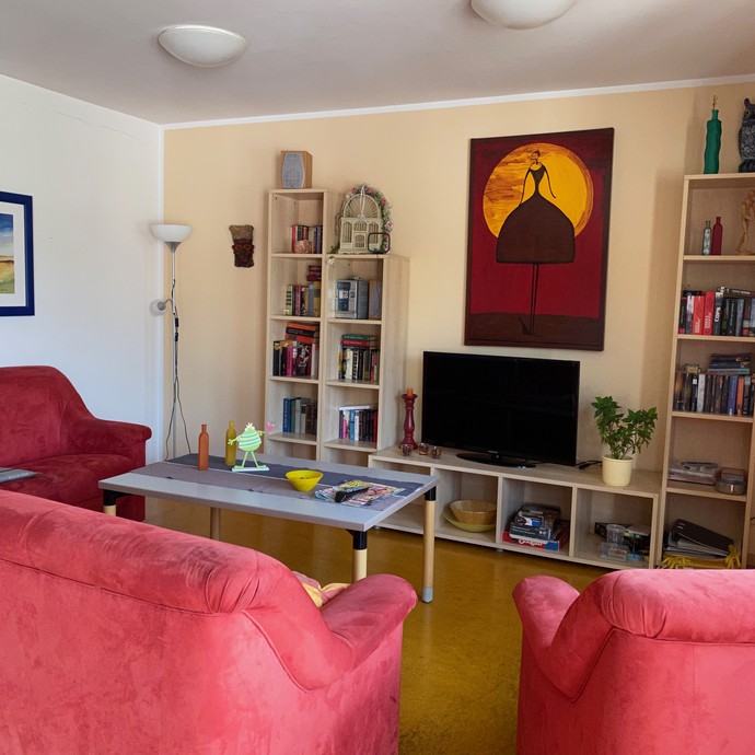 Blick in ein Wohnzimmer mit roten Sitzmöbeln, hinten eine Schrankwand mit Büchern (öffnet vergrößerte Bildansicht)